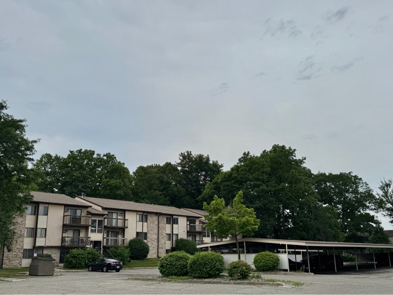 Three-story Kaynorth apartment building with balconies and stone accents, mature trees, and the covered carport parking structure